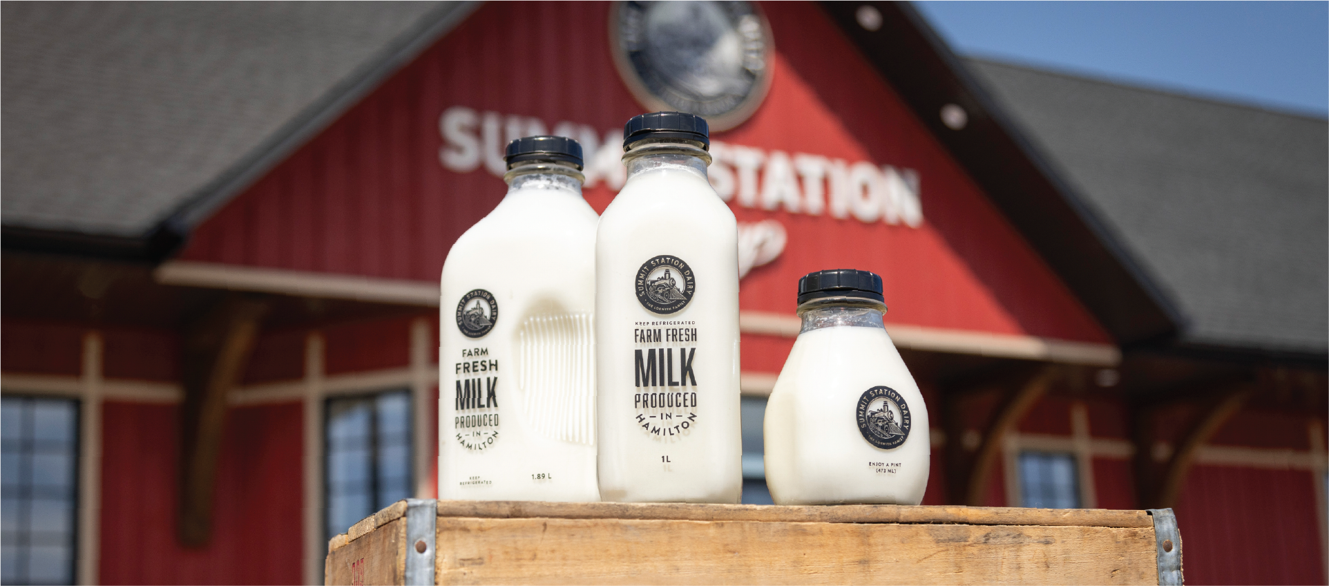 1.89L, 1L, and 473ml bottles of whole milk sitting on a milk crate in front of the red Summit Station on-farm store